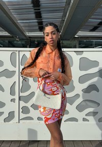 Camicia trasparente arancione, mini gonna a fiori nei colori rosa, rosso e bianco, con una piccola borsa bianca con logo.