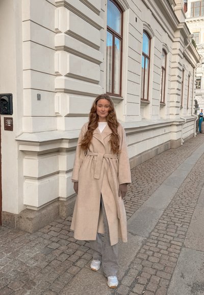 Mujer joven con cabello largo que lleva un abrigo beige con cinturón, guantes, pantalones grises y zapatillas blancas, de pie en una calle empedrada junto a un edificio blanco.