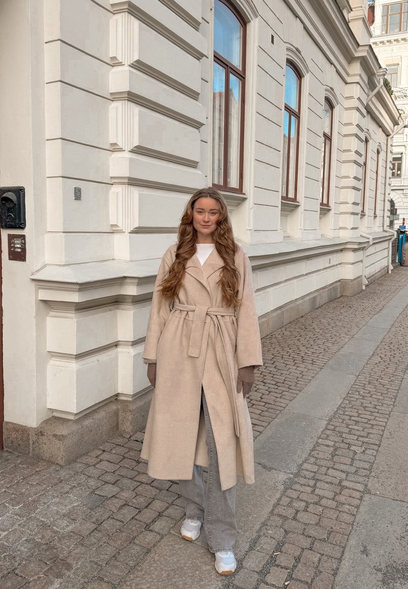 Jeune femme aux longs cheveux portant un manteau beige ceinturé, des gants, un pantalon gris et des baskets blanches, debout dans une rue pavée à côté d'un bâtiment blanc.