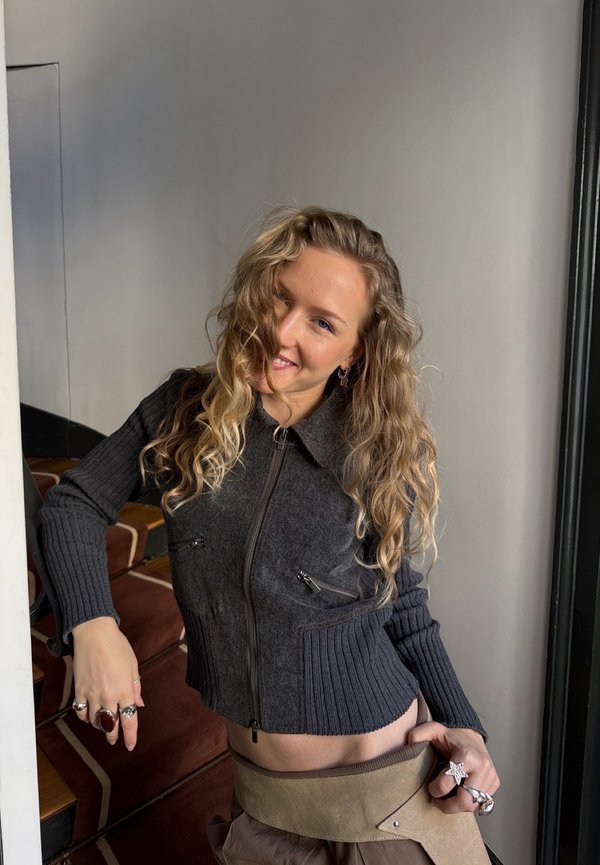 Nuori nainen, jolla on pitkät kiharat hiukset, pukeutuneena tummaan vetoketjulliseen neuleeseen ja useisiin sormuksiin, nojaa sisätiloissa portaikon kaiteeseen.