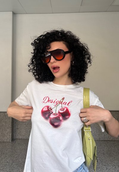 Camiseta blanca con tres gráficos de cerezas rojas con tallo y la palabra "Desigual" en rojo. El accesorio incluye una correa de bolso de color verde lima.