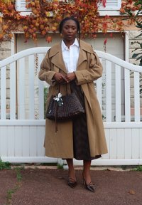 Beżowy trencz na białej koszuli, w zestawieniu z brązową plecioną torebką. Ciemna spódnica w wzory i brązowe szpilki na zakończenie stylizacji.