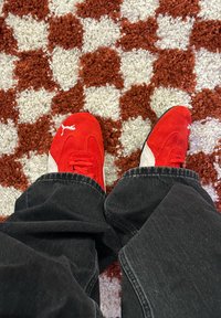 Κόκκινα σουέτ αθλητικά παπούτσια με λευκές λεπτομέρειες και κορδόνια, φορεμένα με μαύρα τζιν παντελόνια, πάνω σε ένα μοτίβο κόκκινο και λευκό shag χαλί.