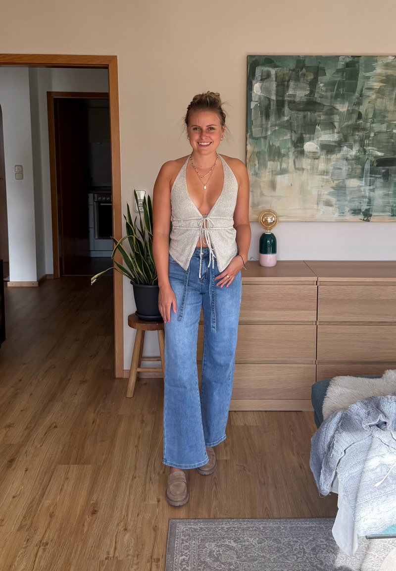 Jeune femme portant un haut dos nu et un jean bleu se tient dans un salon avec un sol en bois, une plante, une commode et une peinture abstraite.