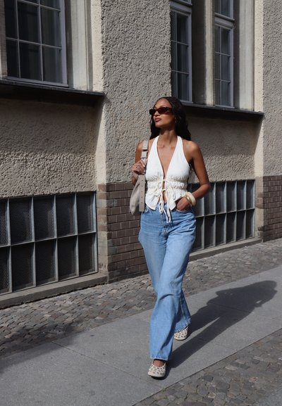 Mujer con gafas de sol, blusa sin mangas blanca, jeans azules y zapatos con tachuelas camina por una calle de adoquines al lado de una pared texturizada.
