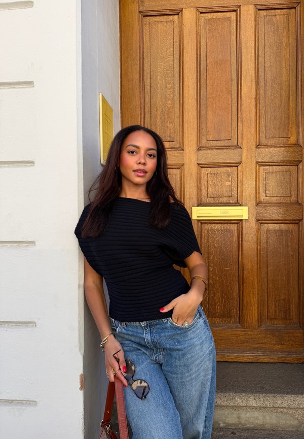 Joven mujer con camiseta negra y vaqueros azules está apoyada en una pared blanca junto a una puerta de madera, sosteniendo unas gafas de sol y un bolso marrón.