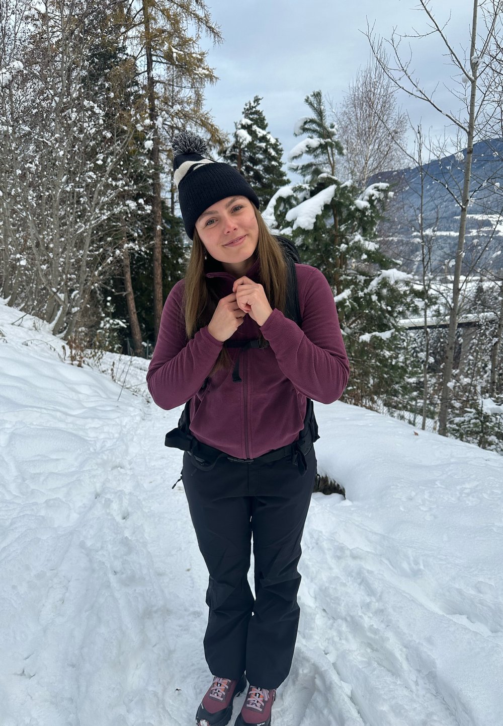 Scène extérieure avec une personne portant un bonnet noir, une veste en polaire violette, un pantalon noir et des chaussures de randonnée, debout sur un sol enneigé.