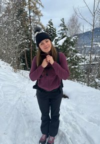 Scène extérieure avec une personne portant un bonnet noir, une veste en polaire violette, un pantalon noir et des chaussures de randonnée, debout sur un sol enneigé.