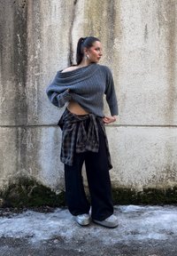 Femme avec une queue de cheval haute portant un pull gris à épaules dénudées, une chemise à carreaux nouée autour de la taille, un pantalon large noir et des chaussures argentées, devant un mur texturé.