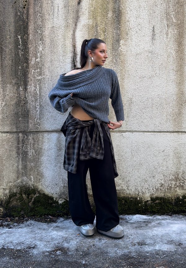 Kvinde med en høj hestehale iført en grå skulderfri sweater, ternet skjorte bundet om livet, vide sorte bukser og sølvfarvede sko foran en struktureret væg.