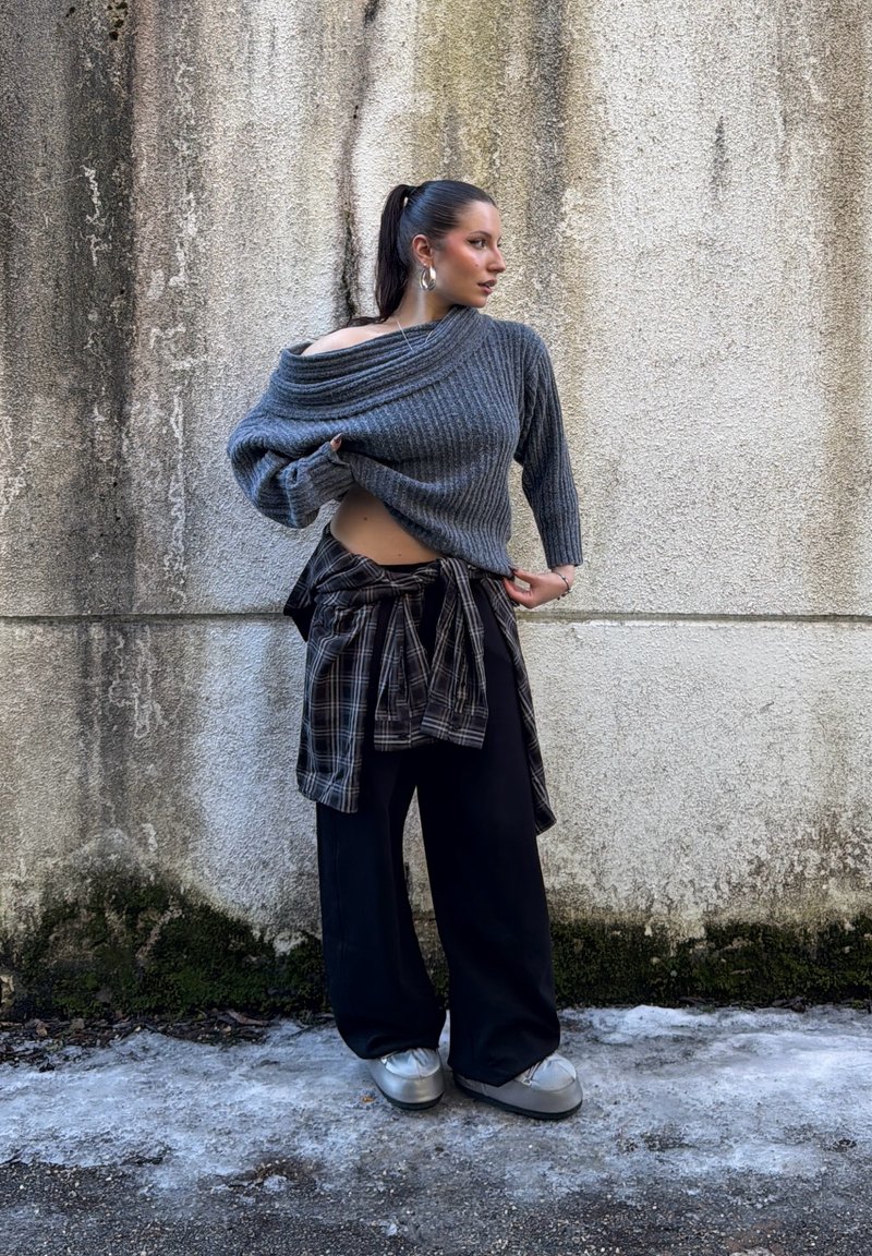 Femme avec une queue de cheval haute portant un pull gris à épaules dénudées, une chemise à carreaux nouée autour de la taille, un pantalon large noir et des chaussures argentées, posant devant un mur texturé.