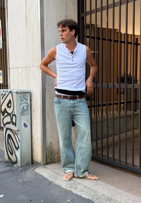 Jeune homme en débardeur blanc, jean ample et tongs appuyé contre un mur près d’une grille métallique dans un cadre urbain.