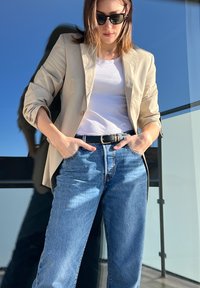 Blazer bege sobre uma camisola branca, cinto escuro e jeans azul claro. O tecido parece suave; o blazer tem um corte descontraído com as mangas arregaçadas.