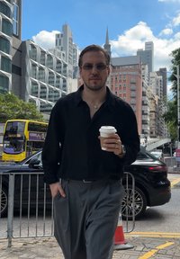 Camisa preta de botão, calças cinzentas, segurando uma chávena de café. Fundo urbano com edifícios modernos, um autocarro amarelo e um carro preto.