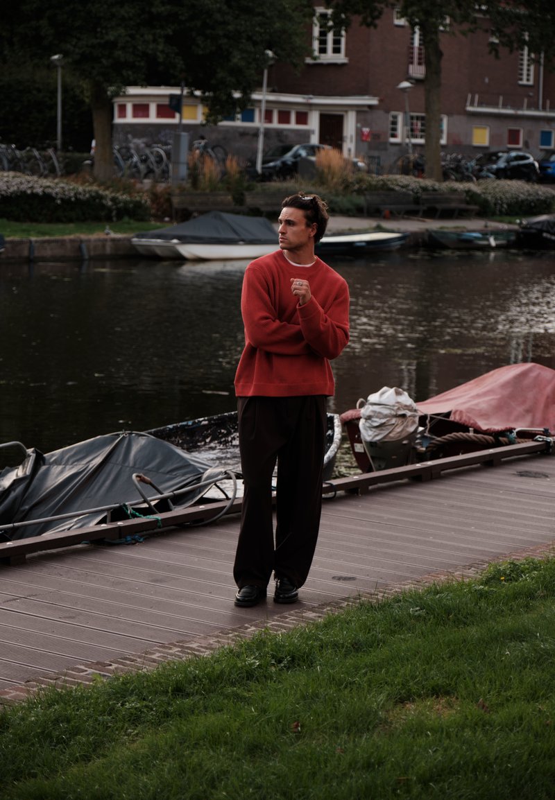 Man in orange sweater standing on a wooden dock by a canal with covered boats and parked cars in the background.