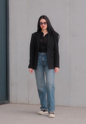Mujer con cabello largo y negro, usando gafas de sol, blazer negro, camisa negra, jeans azules y zapatillas, de pie contra una pared gris.