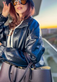 Chaqueta de cuero negro con cierre de cremallera, combinada con una camiseta blanca; sostiene un bolso grande de color marrón oscuro con interior claro, gafas de sol grandes.