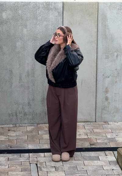 Chaqueta de cuero negra con cuello de piel, pantalones anchos marrones y zapatillas beige. De pie contra una pared texturizada gris, con las manos junto a las orejas.