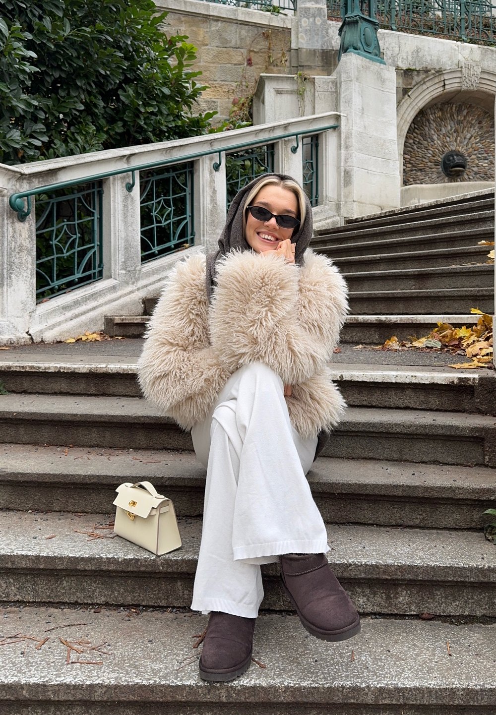 Plysjaktig beige fuskepelsjakke, hvite bukser med vide ben, brune støvler, liten hvit håndveske på trapper med grønt bakteppe.