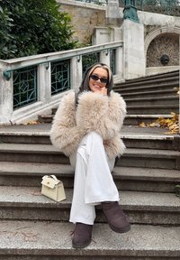 Plysjaktig beige fuskepelsjakke, hvite bukser med vide ben, brune støvler, liten hvit håndveske på trapper med grønt bakteppe.