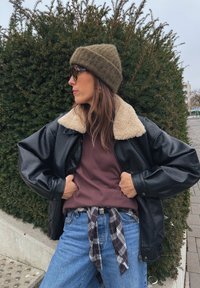 Crna kožna jakna s bež ovčjom ovratnikom, bordo pulover, plave traperice i zelena pletena kapa; prugasta kravata oko struka.