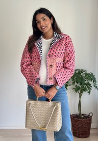 Gesteppte rosa Jacke mit Blumenmuster, blauem Karokragen und braunen Knöpfen, kombiniert mit einer gewebten beigen Handtasche und blauen Jeans.