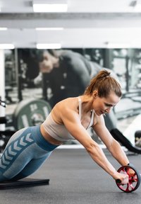 Kvinna i träningskläder som använder en ab roller på en matta, sträcker ut armarna framåt i ett gym med vikter och utrustning i bakgrunden.