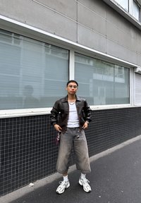 Brązowa skórzana kurtka typu bomber, biały top na ramiączkach, szare szerokie spodnie, białe trampki z akcentami oraz czerwona bandana widoczna na pasku spodni.