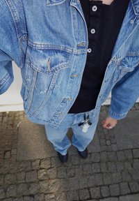 Denim jacket with faded pale blue colour and front pockets, layered over a black henley shirt; pale blue jeans with a loose fit and dark shoes.