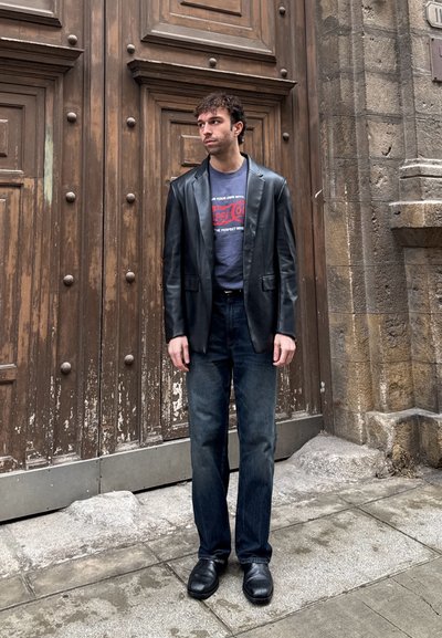Hombre con blazer de cuero negro, camiseta gráfica, vaqueros oscuros y zapatos negros, de pie frente a una gran puerta de madera y una pared de piedra.