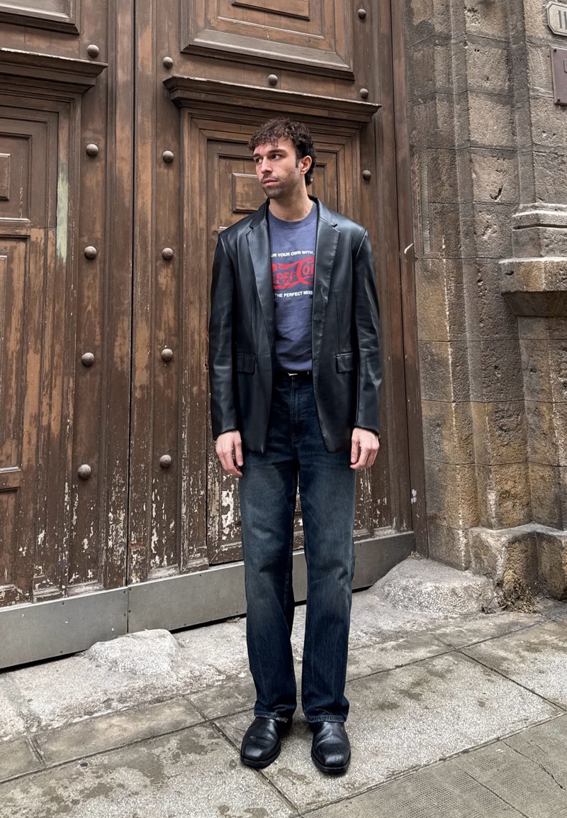 Homme portant un blazer en cuir noir, un t-shirt graphique, un jean foncé et des chaussures noires, debout devant une grande porte en bois et un mur en pierre.