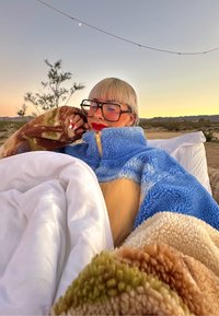 Veste zippée en polaire bleu et beige avec une texture pelucheuse ; manches à motifs marron et vert. Literie blanche et douce à proximité.