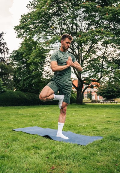 Camisa deportiva verde de manga corta y pantalones cortos a juego. De pie sobre una pierna en una esterilla de yoga azul en la hierba, manos en posición de oración.