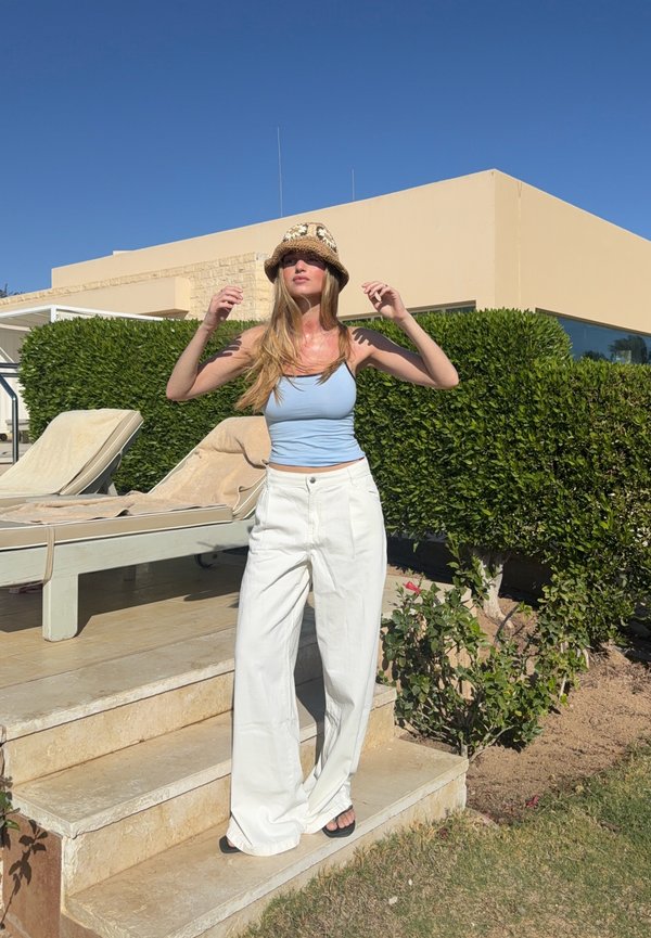 Ženska v širokih belih hlačah, svetlo modri majici brez rokavov in vzorčastem klobuku stoji na kamnitih stopnicah blizu ležalnikov in obrezanih grmičev pod jasnim nebom.