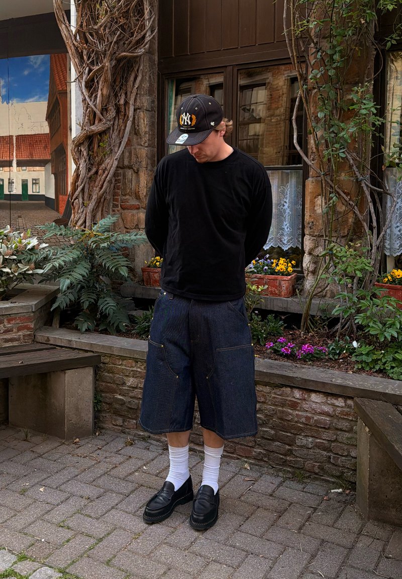 Homme en chemise noire à manches longues, short en jean foncé oversize, chaussettes blanches, mocassins noirs et casquette de baseball noire, debout tête baissée près d’un mur de jardin en briques.