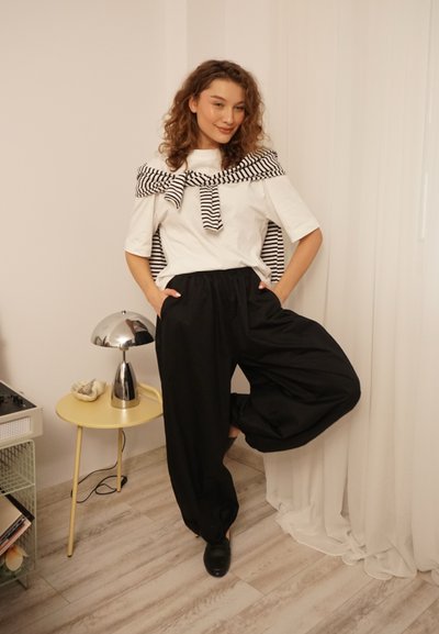 Camiseta blanca con una banda envuelta a rayas en blanco y negro alrededor de los hombros, combinada con pantalones negros de pierna ancha; zapatos negros brillantes y una lámpara plateada.