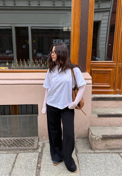 Mujer con gafas que lleva una camisa blanca de manga larga y pantalones anchos negros, de pie en una acera de piedra cerca de una puerta y ventana de madera.