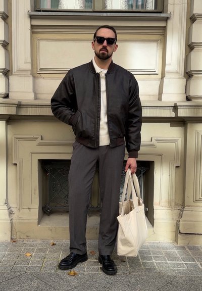 Hombre vestido con una chaqueta bomber negra, pantalones grises, zapatos negros y gafas de sol, sosteniendo una bolsa de tela beige mientras está de pie en una acera de baldosas.