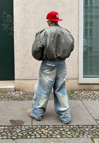 Chaqueta bomber verde oliva, pantalones vaqueros claros y oversized de mezclilla con desgastes y rotos, zapatos beige, gorra roja con logo blanco de NY, de pie sobre adoquines.