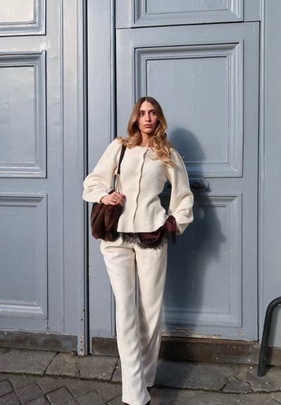 Mujer con cabello largo y rubio, vestida con un suéter y pantalones color crema, sosteniendo un bolso marrón, de pie frente a una gran puerta azul con paneles.
