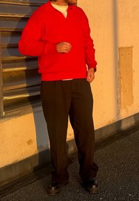 Κόκκινο πλεκτό πουλόβερ με ρίγες και πόλο γιακά, συνδυασμένο με μαύρο παντελόνι και σκούρα παπούτσια. Στεκόμαστε μπροστά σε έναν υφασμάτινο τοίχο κάτω από το απαλό ηλιακό φως.