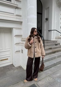 Femme portant un manteau en fourrure beige, un pantalon large foncé et des lunettes de soleil, debout sur un trottoir gris près des marches d'un bâtiment blanc, tenant un sac à main marron.
