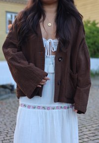 Bruine, getextureerde stoffen jas met wijde mouwen en grote knopen, gedragen over een witte jurk met bloemrijke borduursels bij de taille.