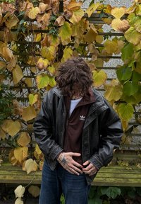Sort læderjakke over en brun lynlås sweatshirt med et hvidt logo, parret med blå jeans. Omgivet af efterårsblade.