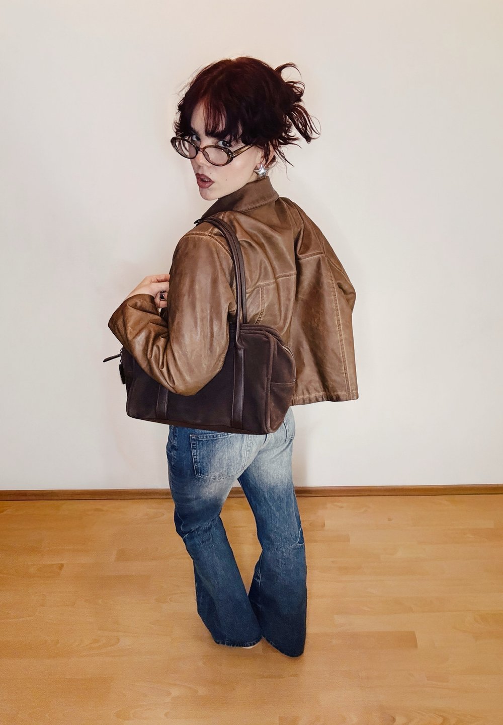 Veste en cuir marron au design court, sac à bandoulière marron et jean évasé bleu. Texture lisse, détails minimaux et fond neutre.