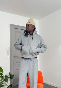 Mand i grå hættetrøje og joggingbukser, iført en beige hue, står indendørs ved en grå dør og en orange stol, og kigger til siden.