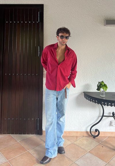 Camisa roja de botones, con el cuello desabrochado, jeans azul claro y botas de vaquero negras, con gafas de sol; texturas suaves y corte relajado.