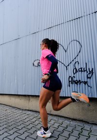 Donna in abbigliamento da corsa rosa e blu che corre davanti a un muro di metallo ondulato con un cuore di graffiti e testo.