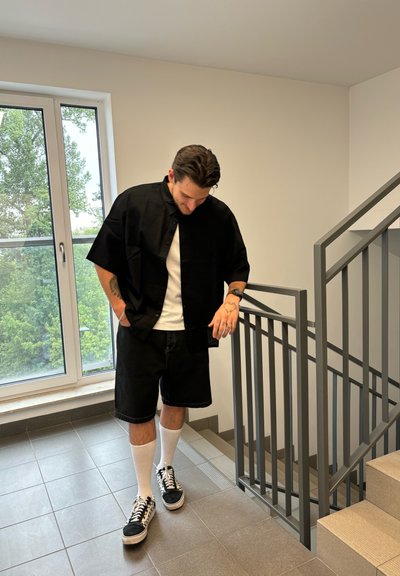 Hombre con tatuajes vestido con una camiseta negra, pantalones cortos, calcetines blancos y zapatillas de deporte, de pie junto a la barandilla de una escalera en un espacio interior alicatado cerca de una ventana.