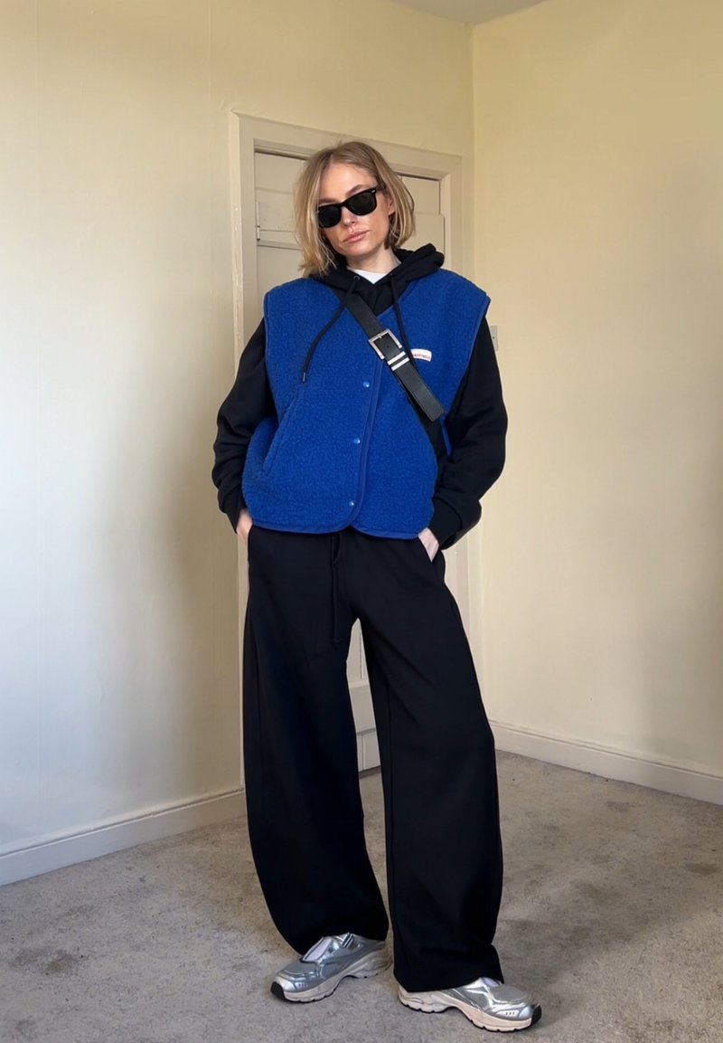 Persona con felpa nera con cappuccio, gilet in pile blu senza maniche, pantaloni neri larghi, sneaker argento e occhiali da sole neri in piedi all'interno.